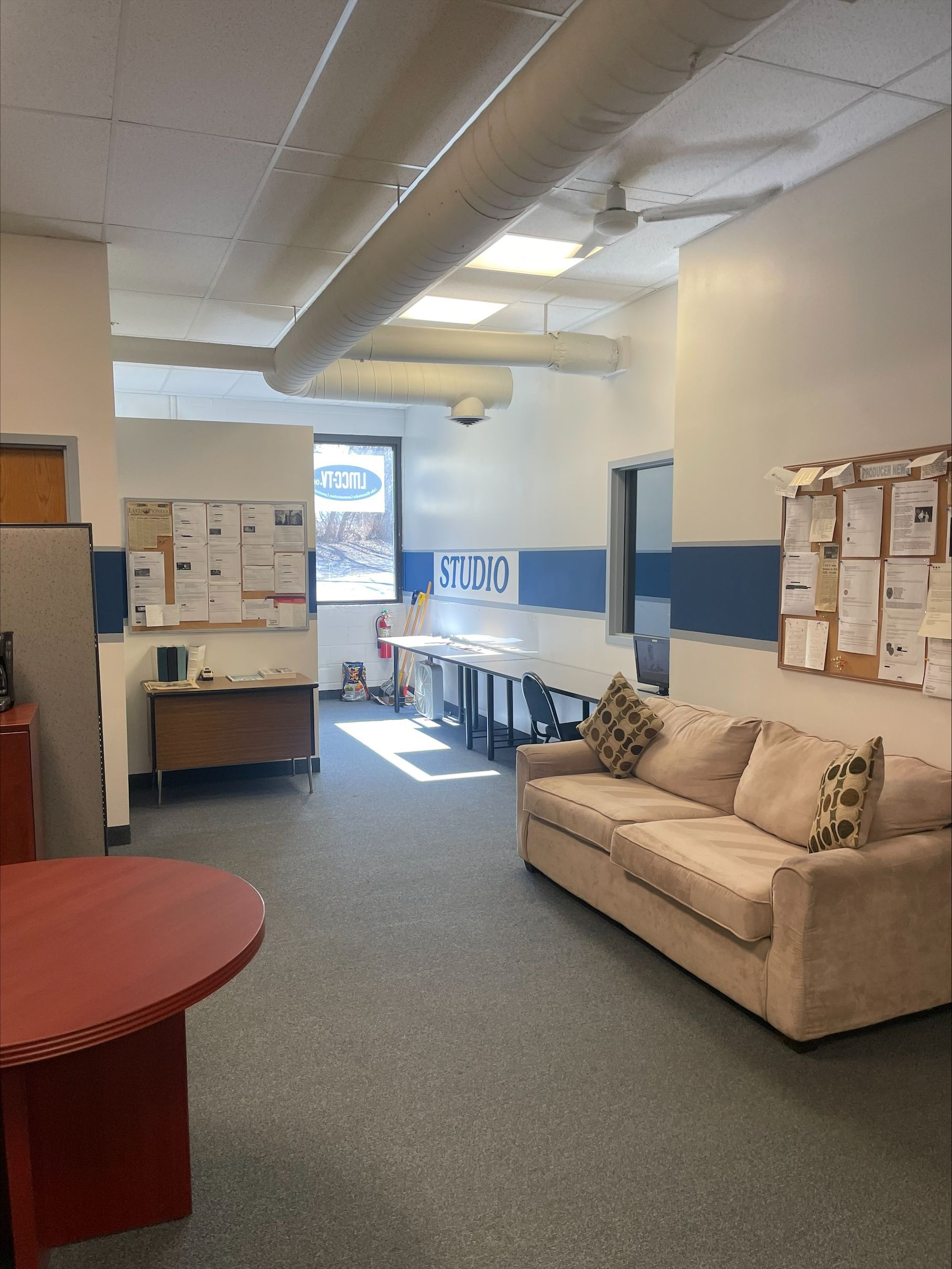 Office with a red table, sofa, desks, and bulletin boards. Sunlight streams in from a window.