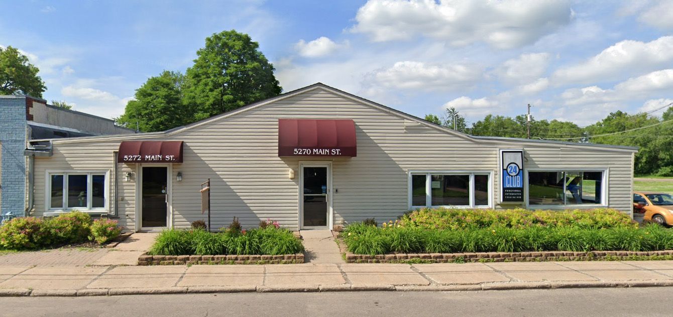 A beige building with maroon awnings and two doors. Bushes line the front; a blue sky is overhead.