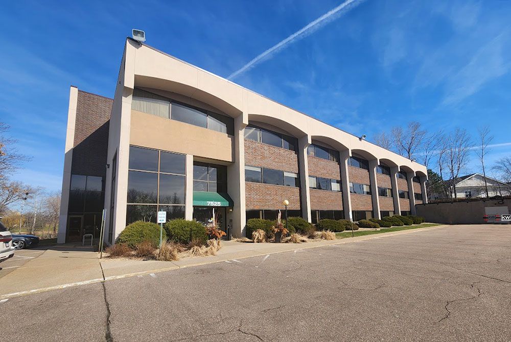 Two-story office building with glass windows, tan facade, and a dark brown side under a blue sky.