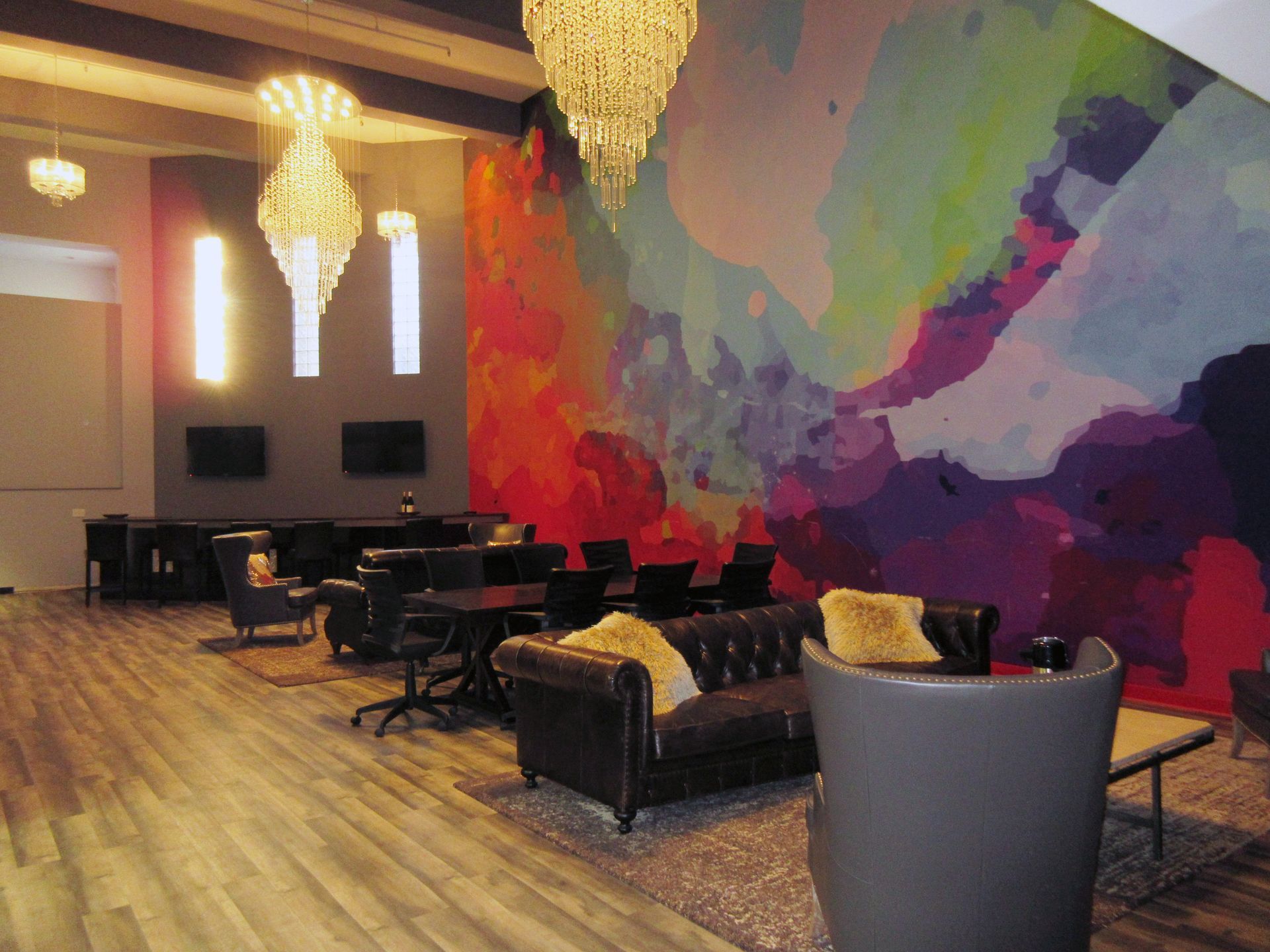 A lounge with hardwood floors, leather seating, and a colorful mural. Chandeliers hang from the ceiling.