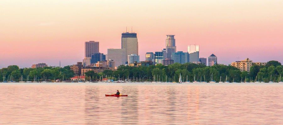 Kayaker in a red boat on water with Minneapolis city skyline in the background at sunset.