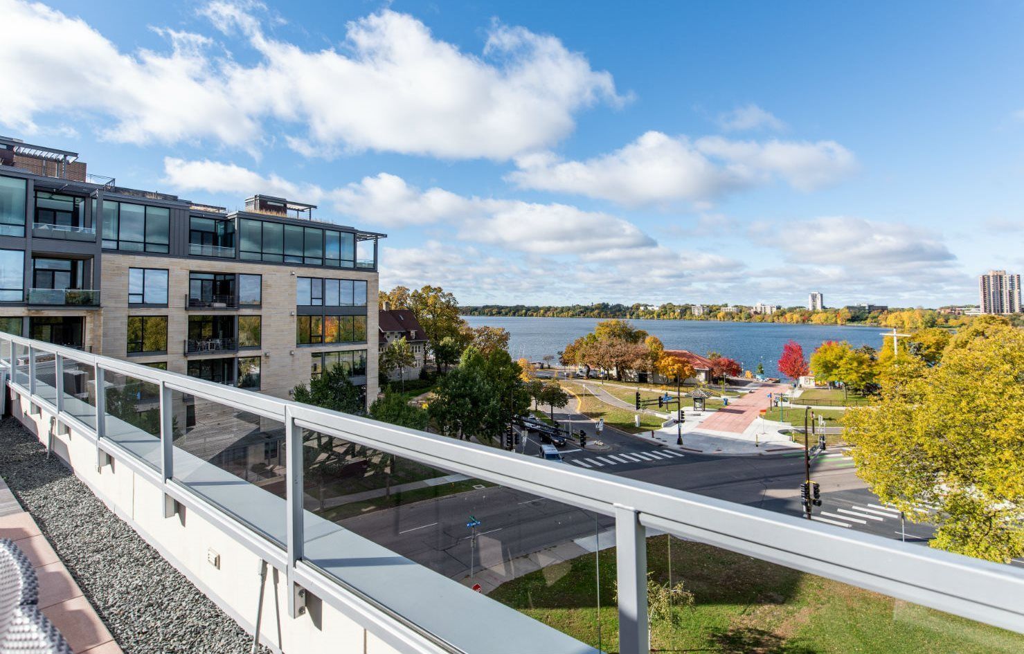 View from a balcony overlooking a street, park, and lake on a sunny day, with buildings and trees.