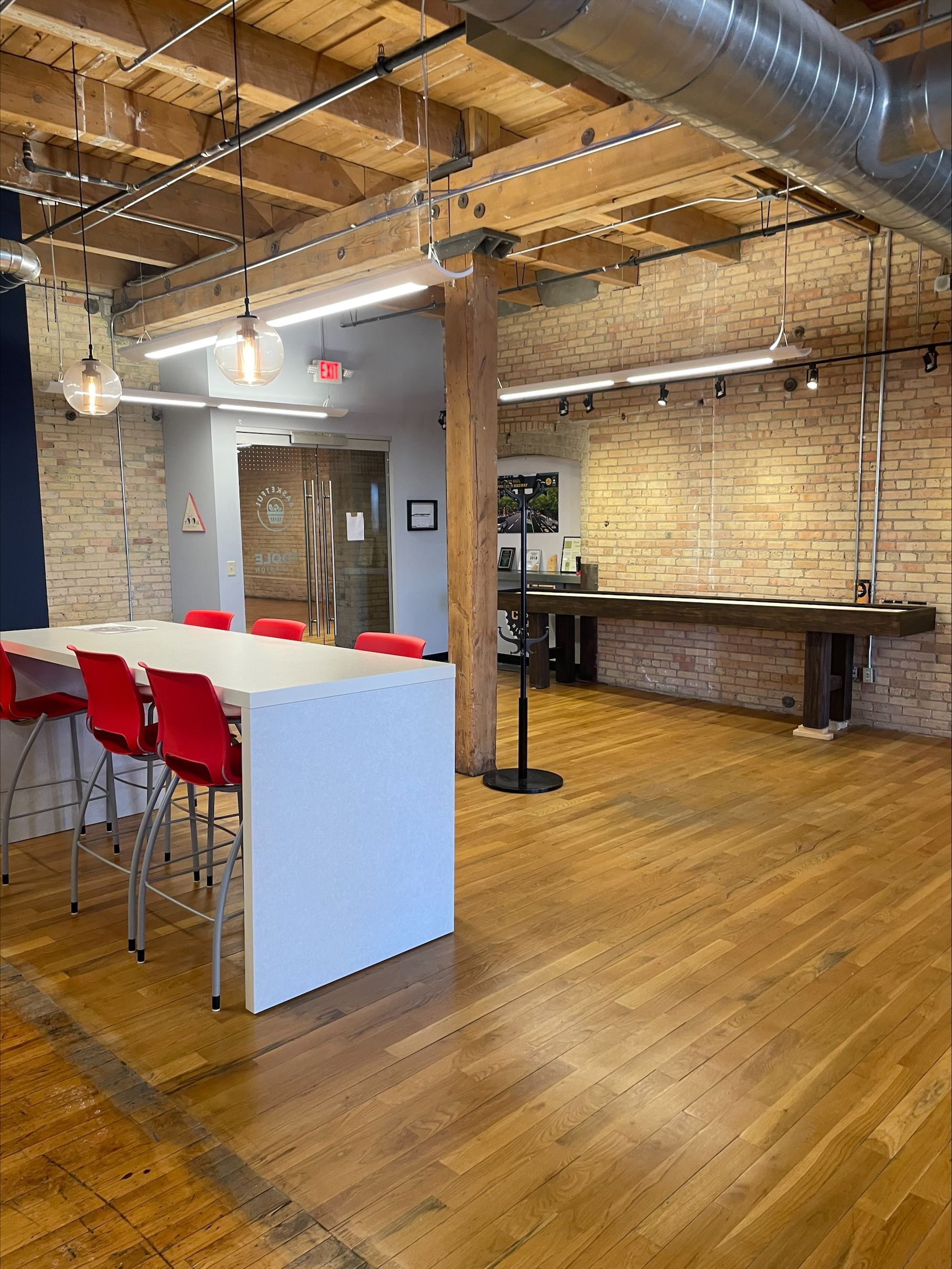 Open office space with wooden floors, exposed brick and beams, white table with red chairs.
