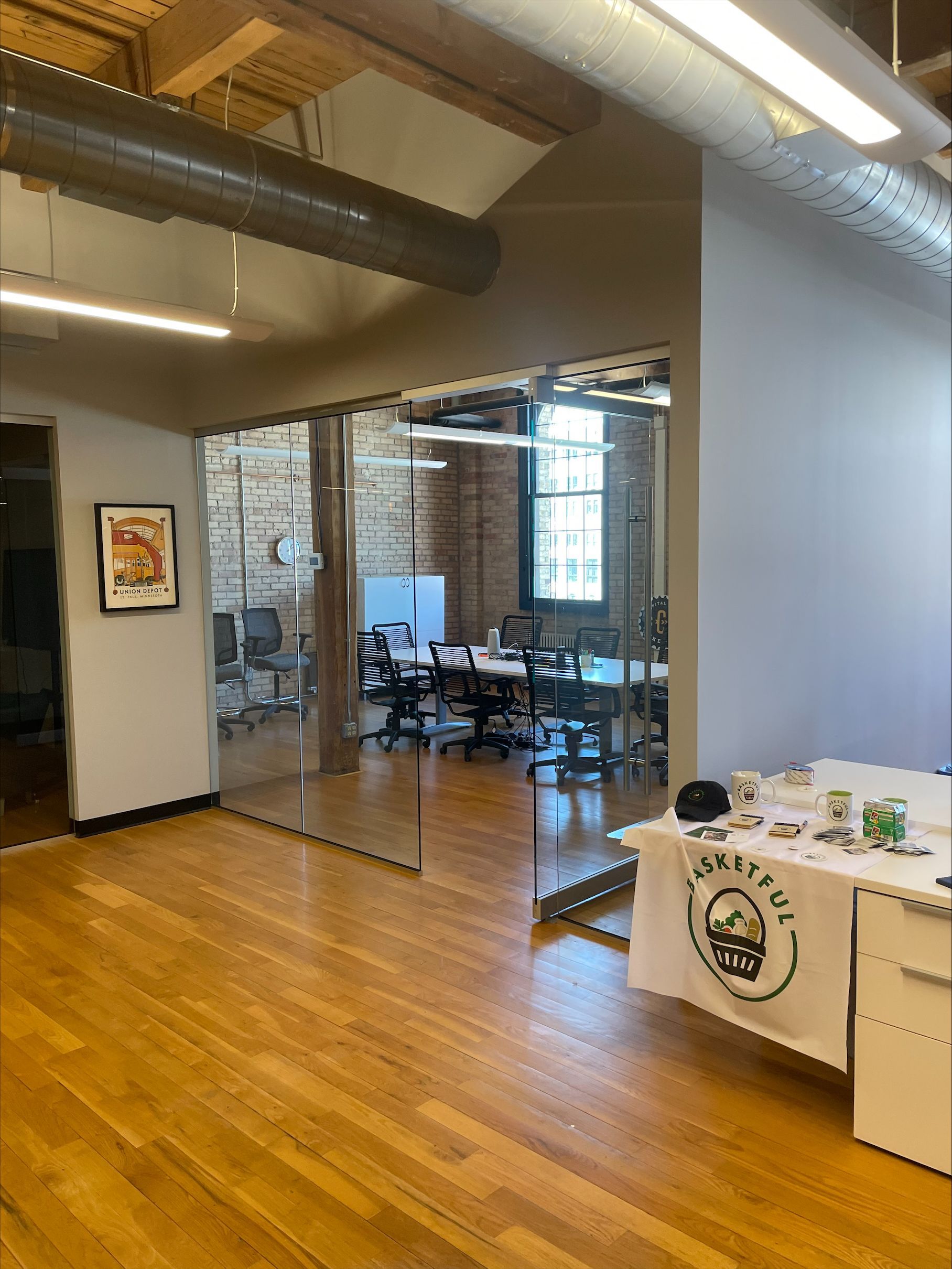 Wooden office floor with a glass-walled conference room, a desk with a logo, and exposed ductwork on the ceiling.