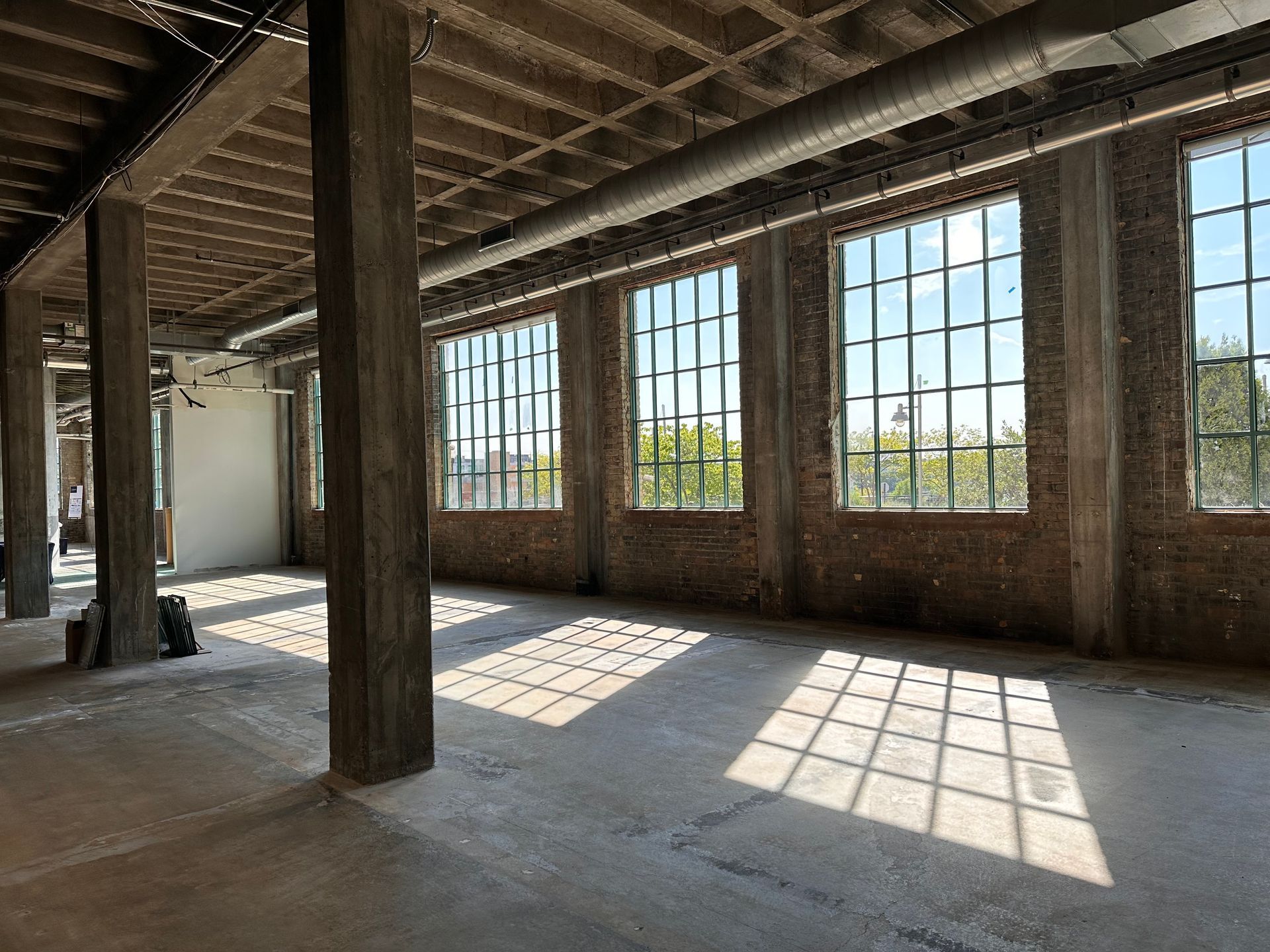 An empty industrial space with large windows, brick walls, concrete pillars, and sunlight streaming through.