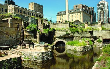Historic ruins with a water channel in the foreground, industrial buildings and modern skyscrapers in the background.