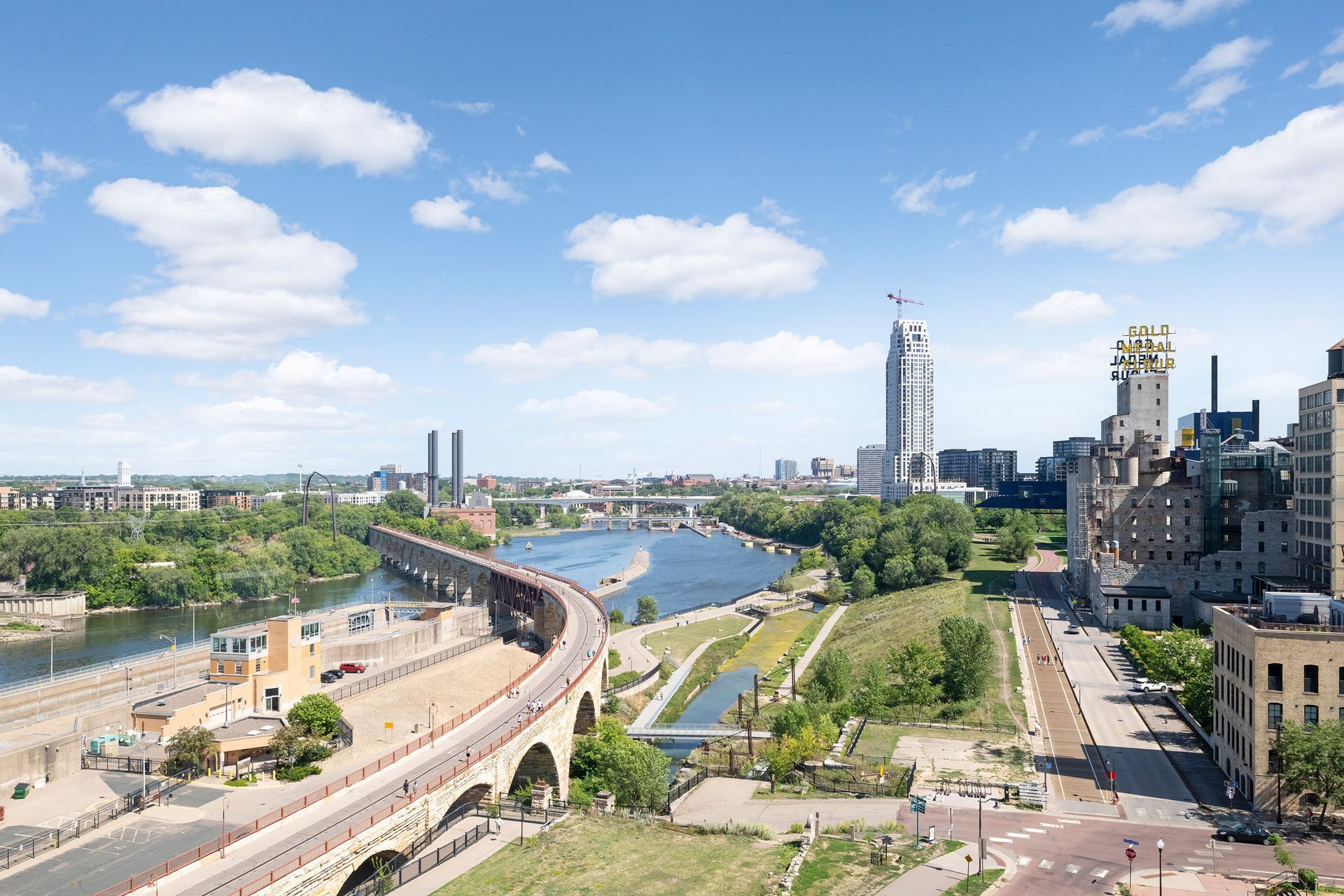 Cityscape overlooking a river, with a bridge, buildings, and blue sky with clouds.