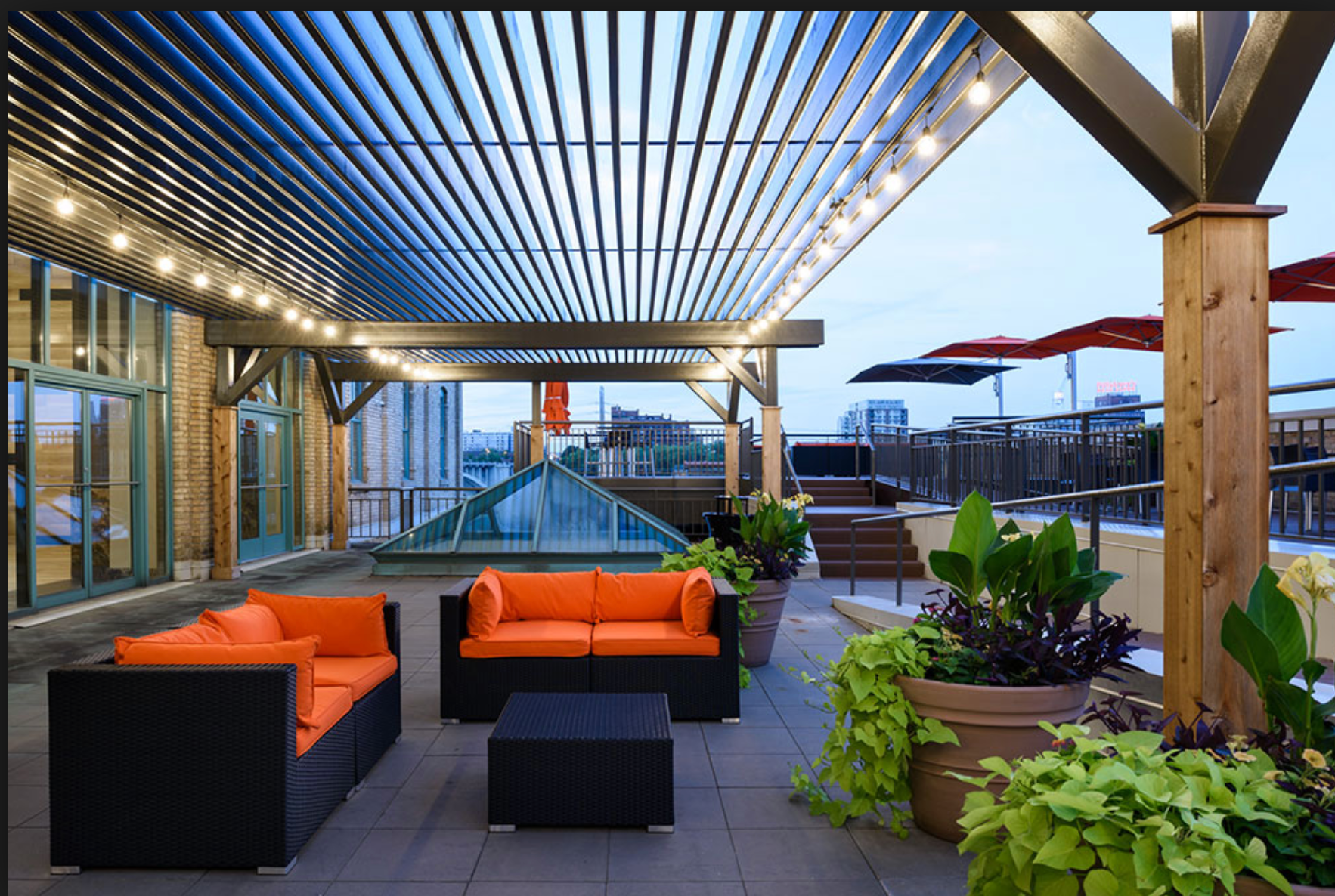 Rooftop patio with orange couches, plants, and string lights. Wooden beams support the overhead structure.