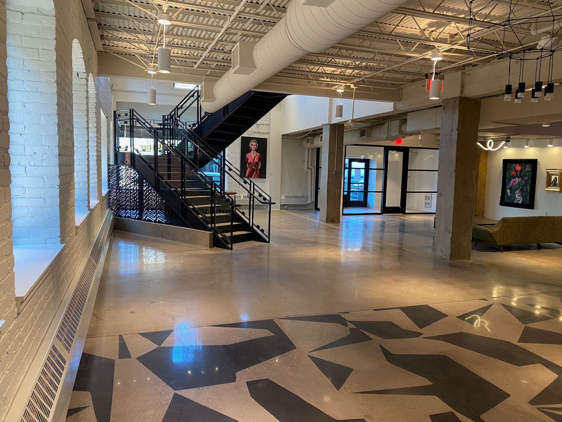 Large open interior with a modern staircase and abstract floor design. White brick walls, dark metal stairs, and artwork.