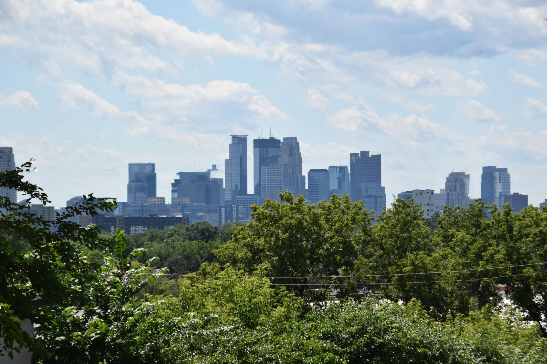 Skyline of Minneapolis, Minnesota with tall buildings against a cloudy sky, trees in the foreground.