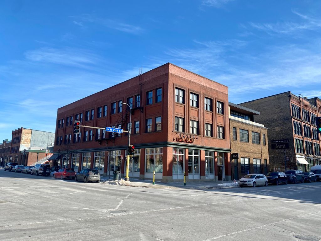 Red brick building on a city corner with storefronts, cars parked on street, blue sky.