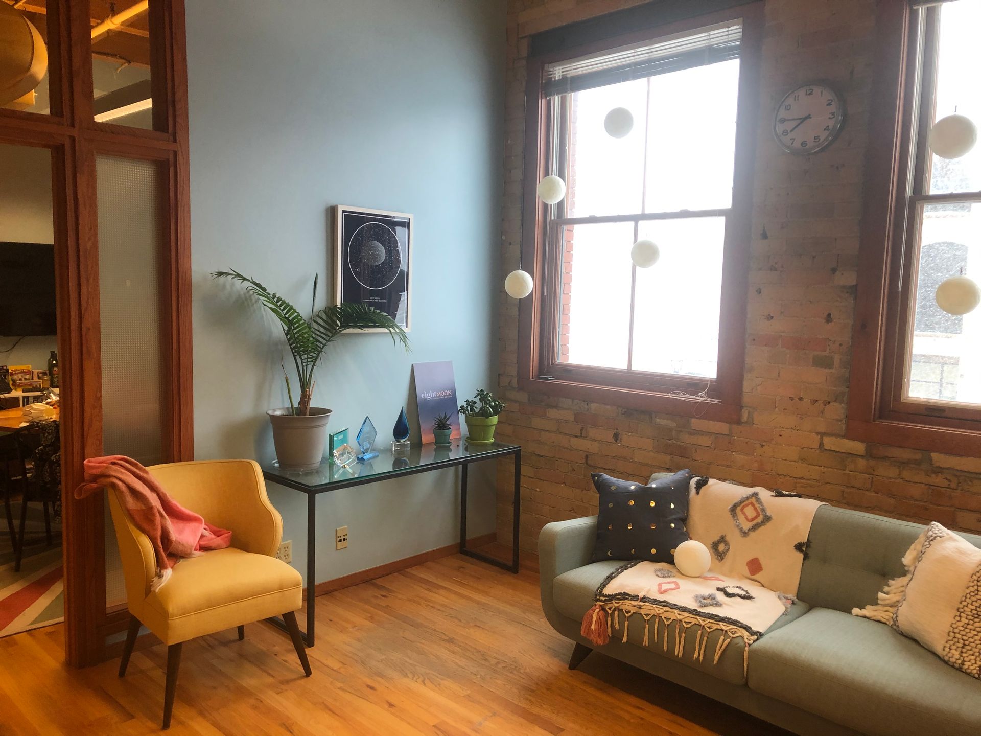 Office area with a blue-green couch, yellow chair, brick wall, and glass-topped table with plants.