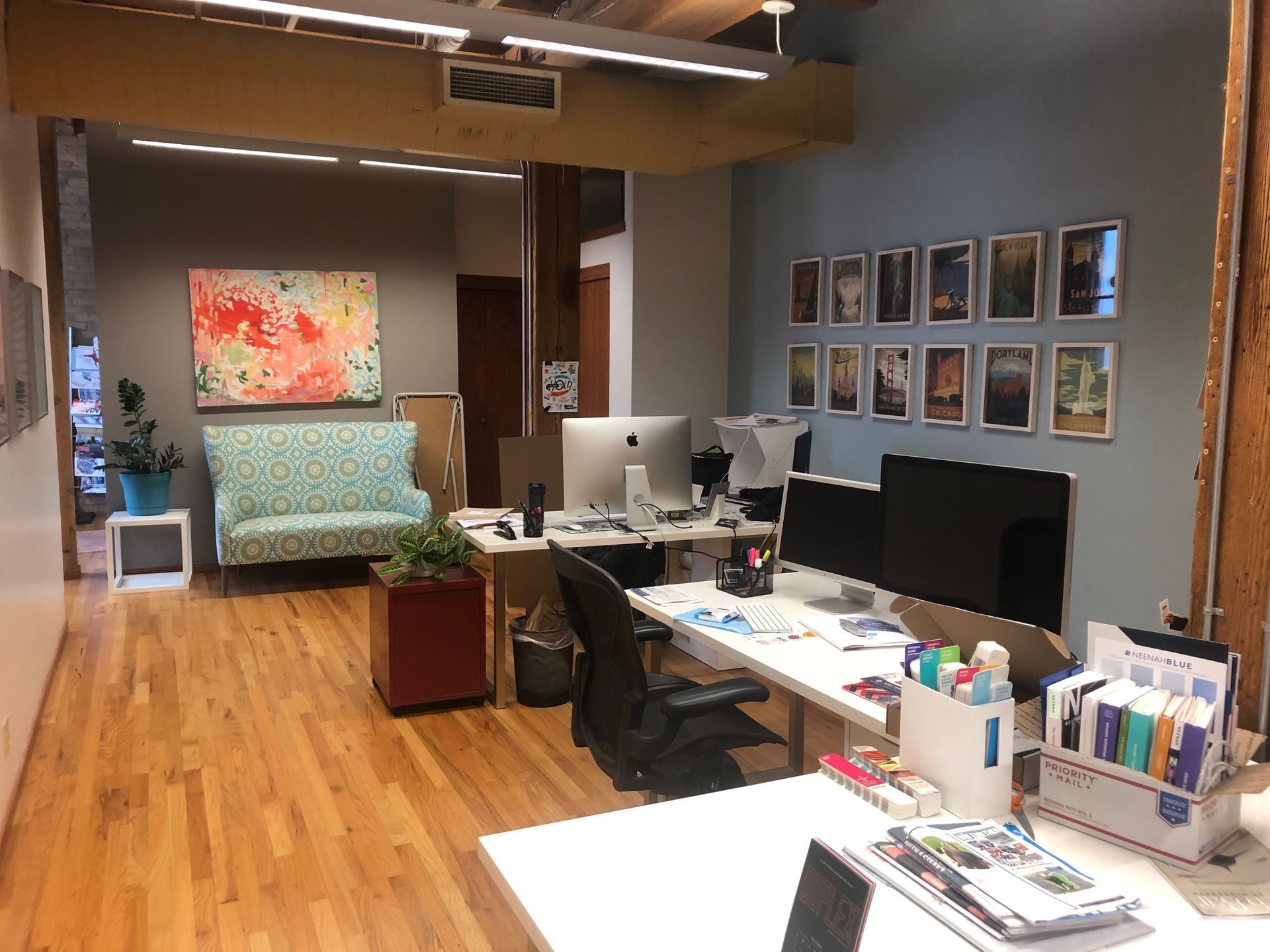 Modern office space with desks, computers, a couch, and framed art on the wall. Wooden floors, and colorful decor.