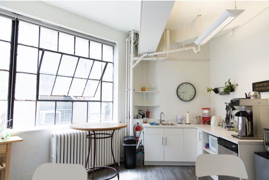 Office space with large windows, small kitchen, and round table. White walls, gray floor.
