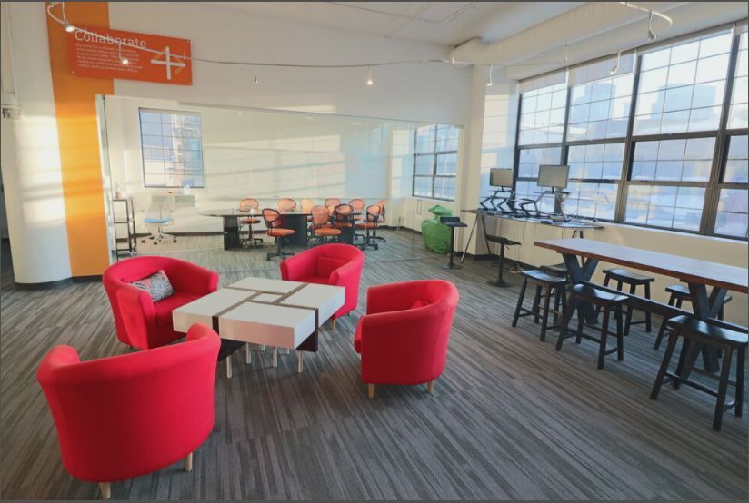 Spacious office area with red seating, a long table with stools, and large windows; natural light.