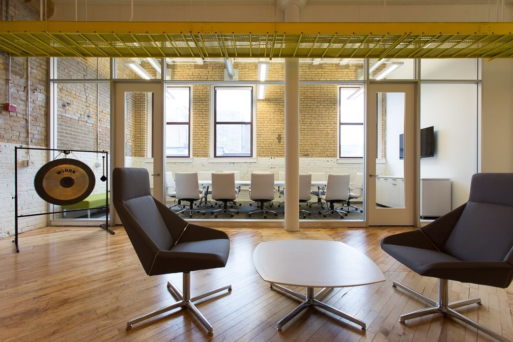 Modern office waiting area with lounge chairs, coffee table, and gong, overlooking a conference room.