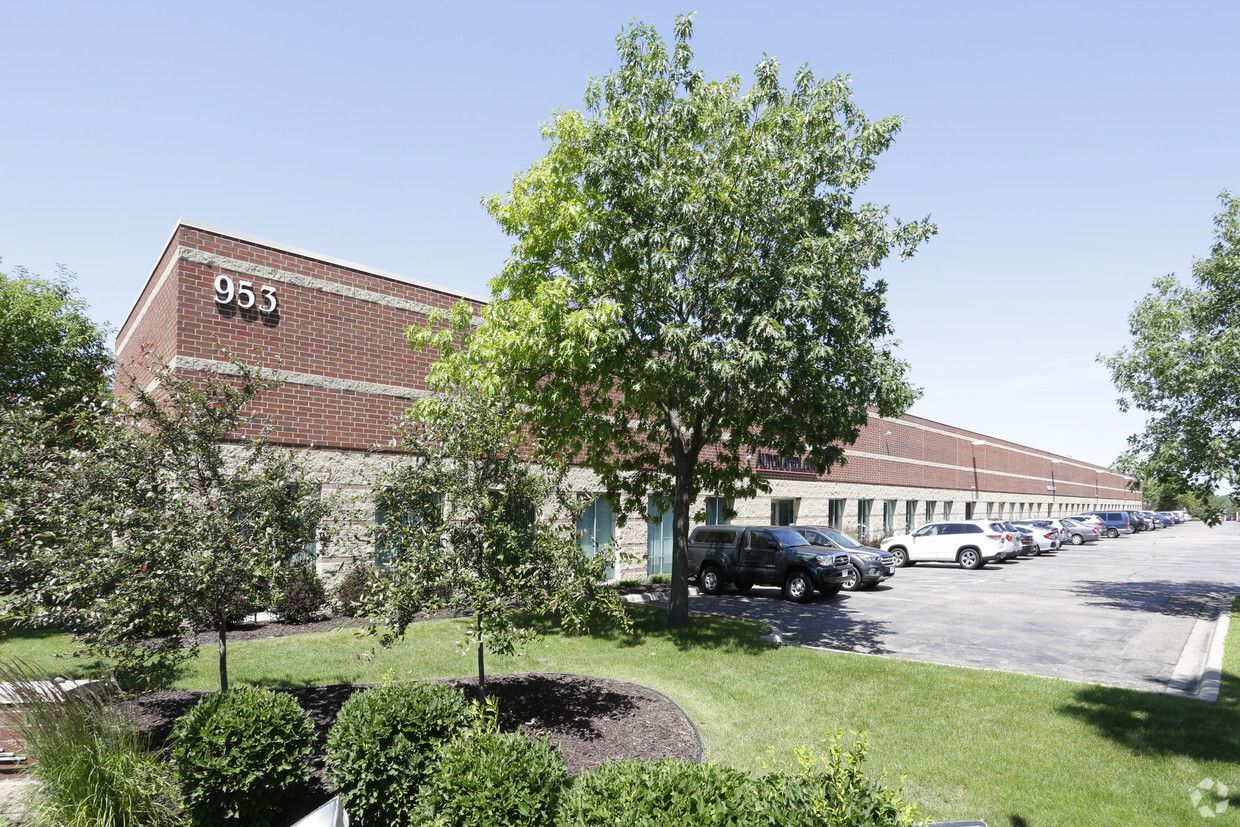 Brick industrial building with parked cars and green landscaping on a sunny day.