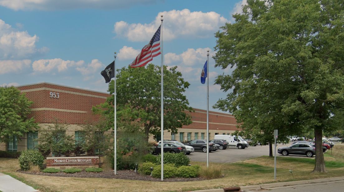 Flags fly in front of a brick building with parked cars and trees on a sunny day.