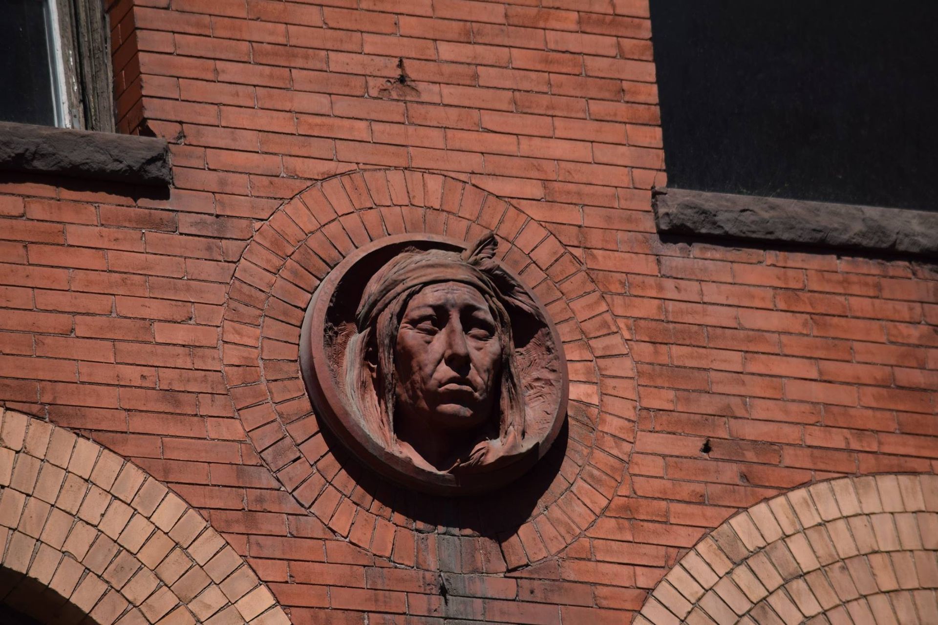 Brick building with carved, circular relief of a person's face, framed by an arched brick pattern.