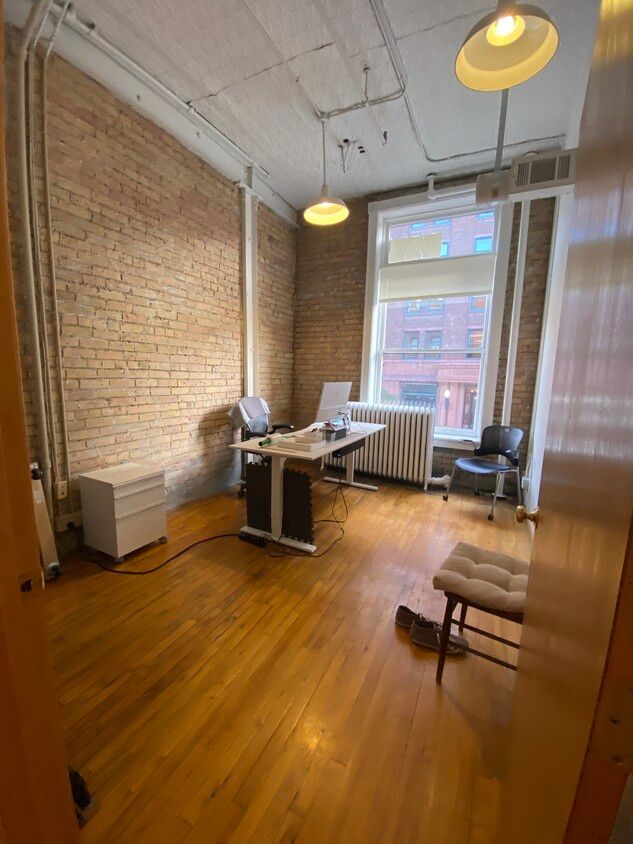 Office with exposed brick walls, wood floor, desk, chairs, and large window. Two pendant lights hang from the ceiling.