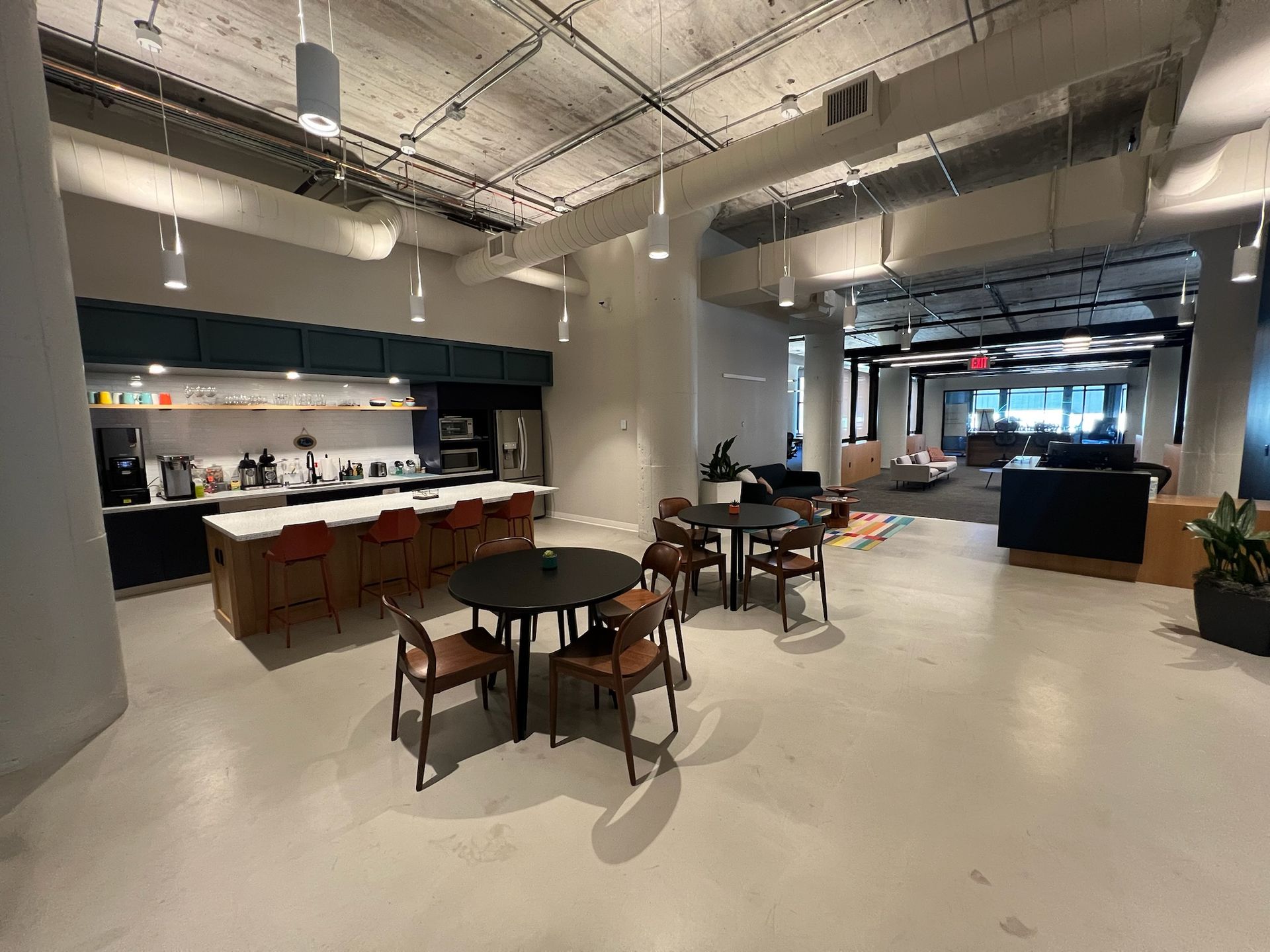 Open-plan office kitchen and seating area with tables, chairs, counters, and a fridge.
