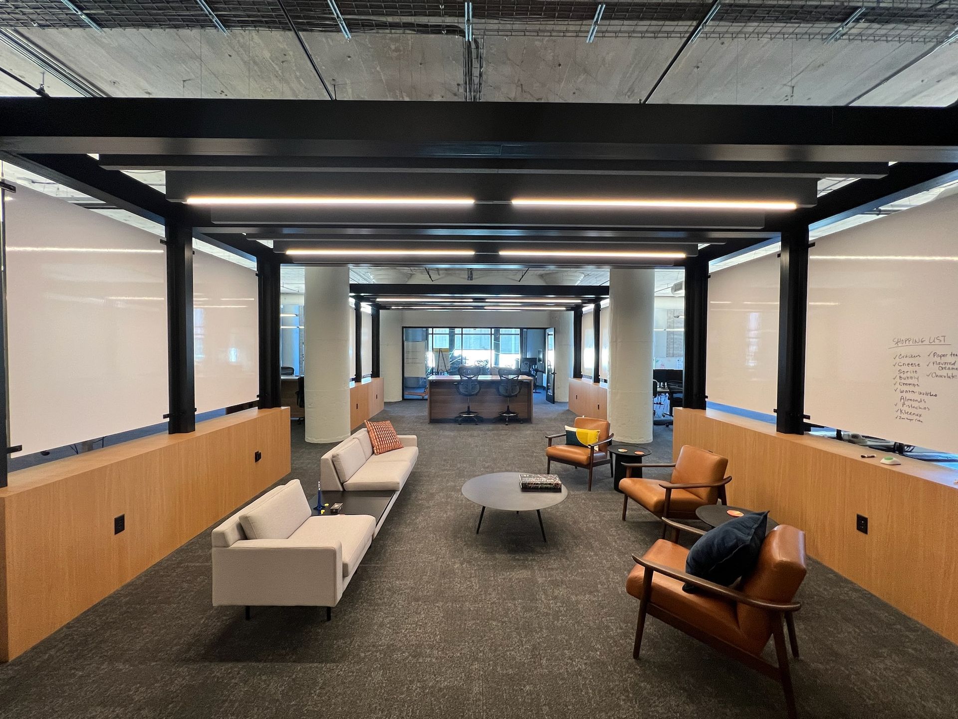Modern office lounge with couches, chairs, and coffee table under a black beam structure.