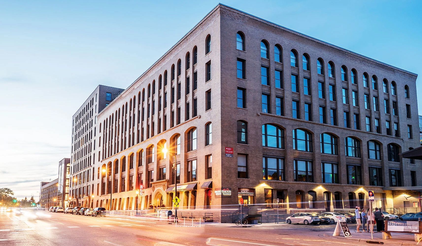 Brick building, street view at dusk, cars parked. People walk on the sidewalk.