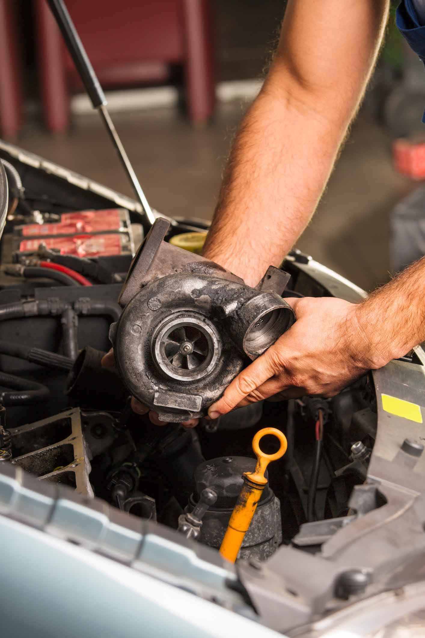 A man is holding a turbocharger under the hood of a car.
