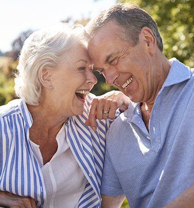 Pareja de ancianos sonrientes, con las caras juntas. Mujer con camisa azul a rayas, hombre con polo. Al aire libre.