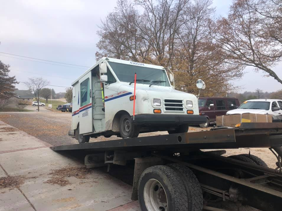 White USPS mail truck on a tow truck, parked on a street with fall foliage.