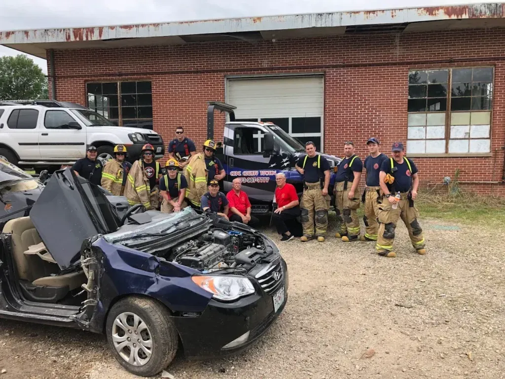 Firefighters pose with a wrecked car in front of a brick building.