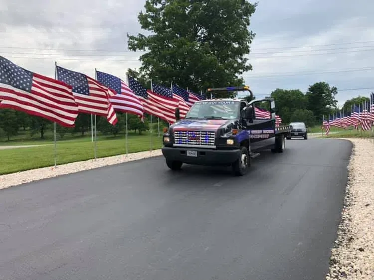 Tow truck driving on a road lined with American flags; cloudy sky, grassy area.