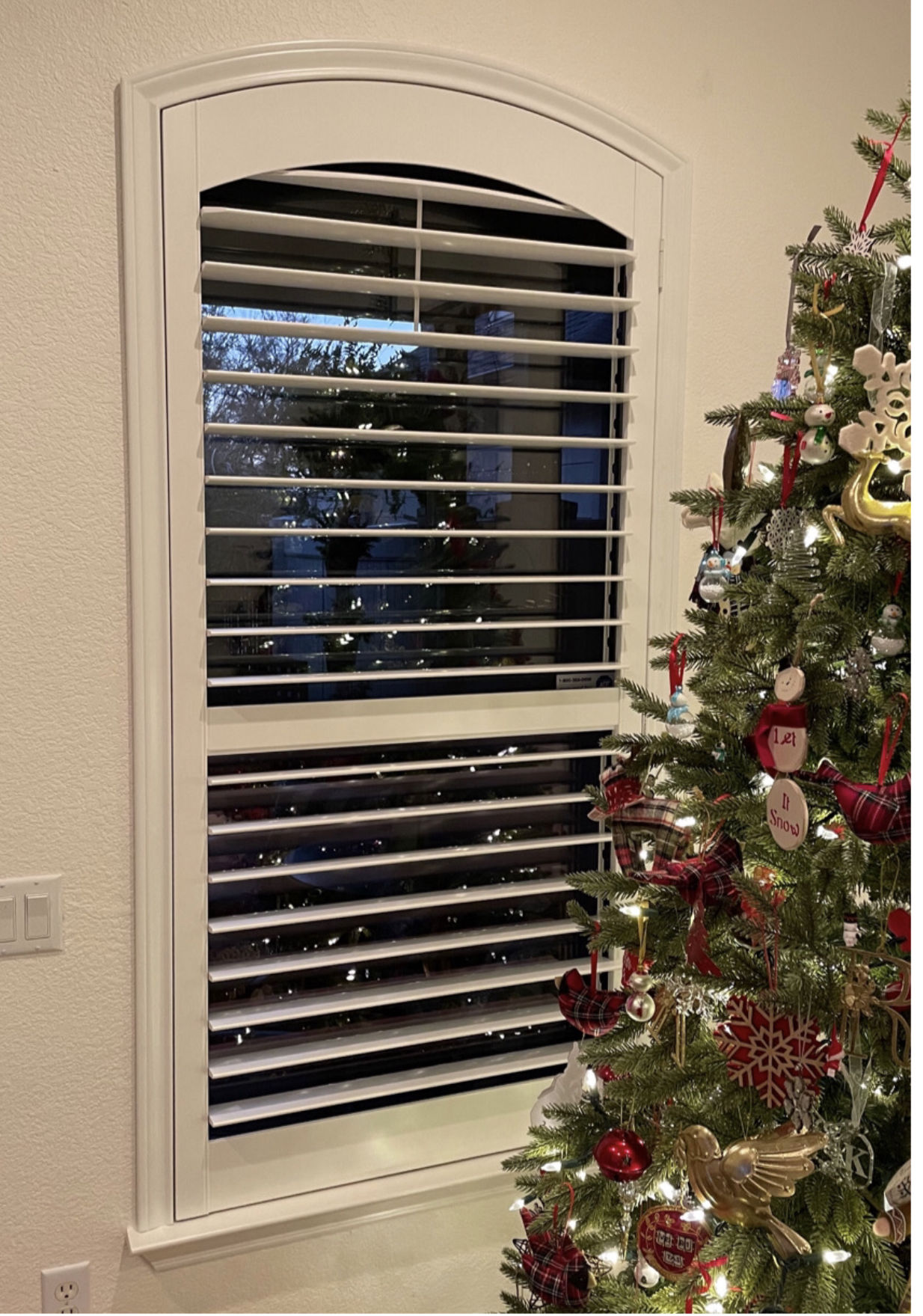A christmas tree is in front of a window with shutters.