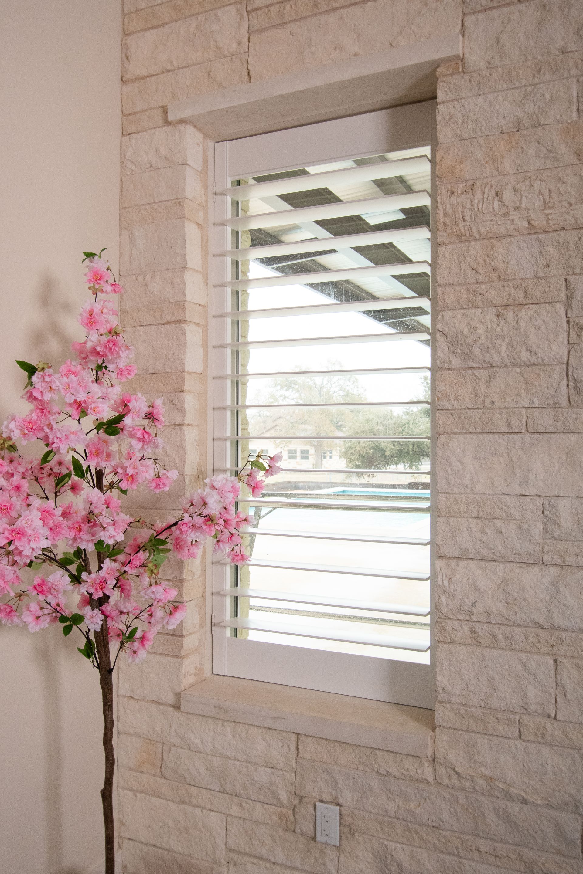 A window with shutters on a brick wall next to a tree with pink flowers.