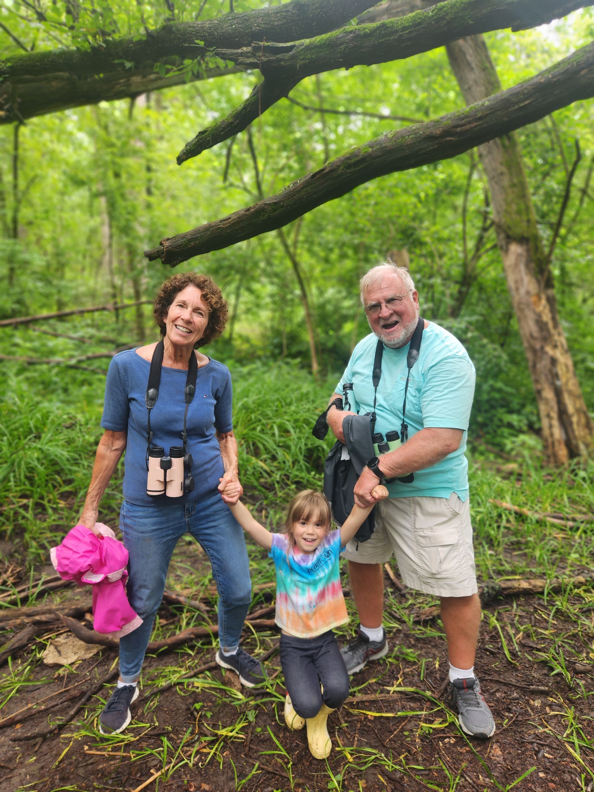 Two adults with binoculars hold hands with a smiling child in the woods.