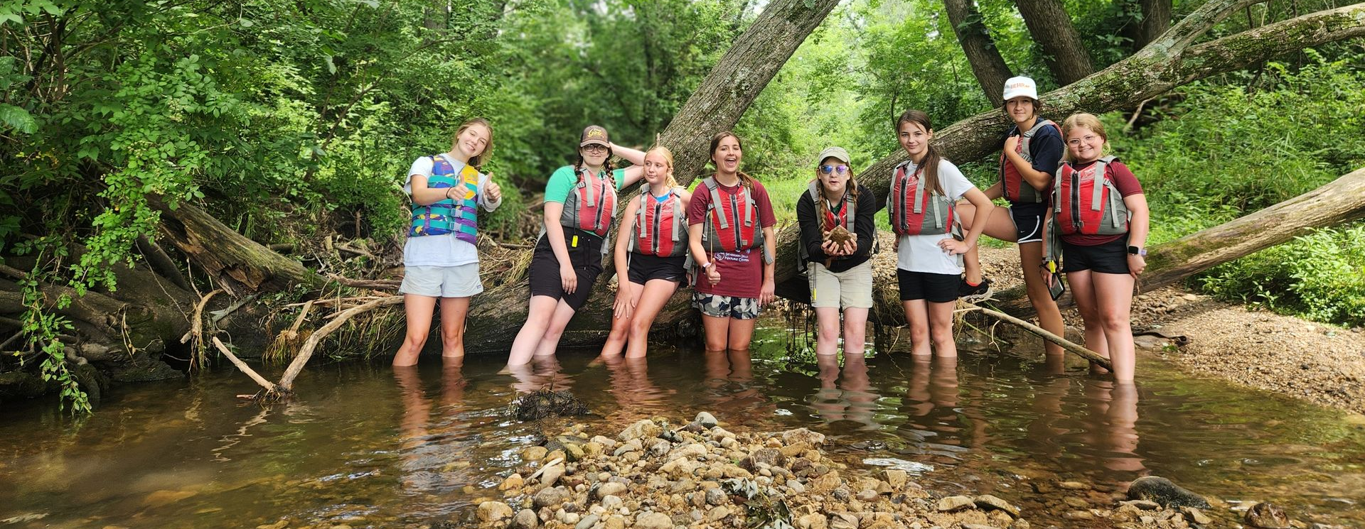 Eight teens standing in a shallow creek, making various poses along a log. The teens are dressed in shorts and t-shirts, all are wearing life jackets.