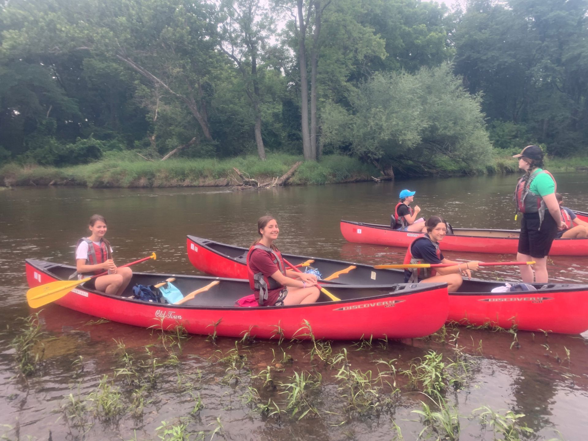 A group of teens sits in three canoes, with a camp leader standing at the front of the middle canoe. They sit in a shallow river wearing life jackets. Three of the teens are smiling for the camera.