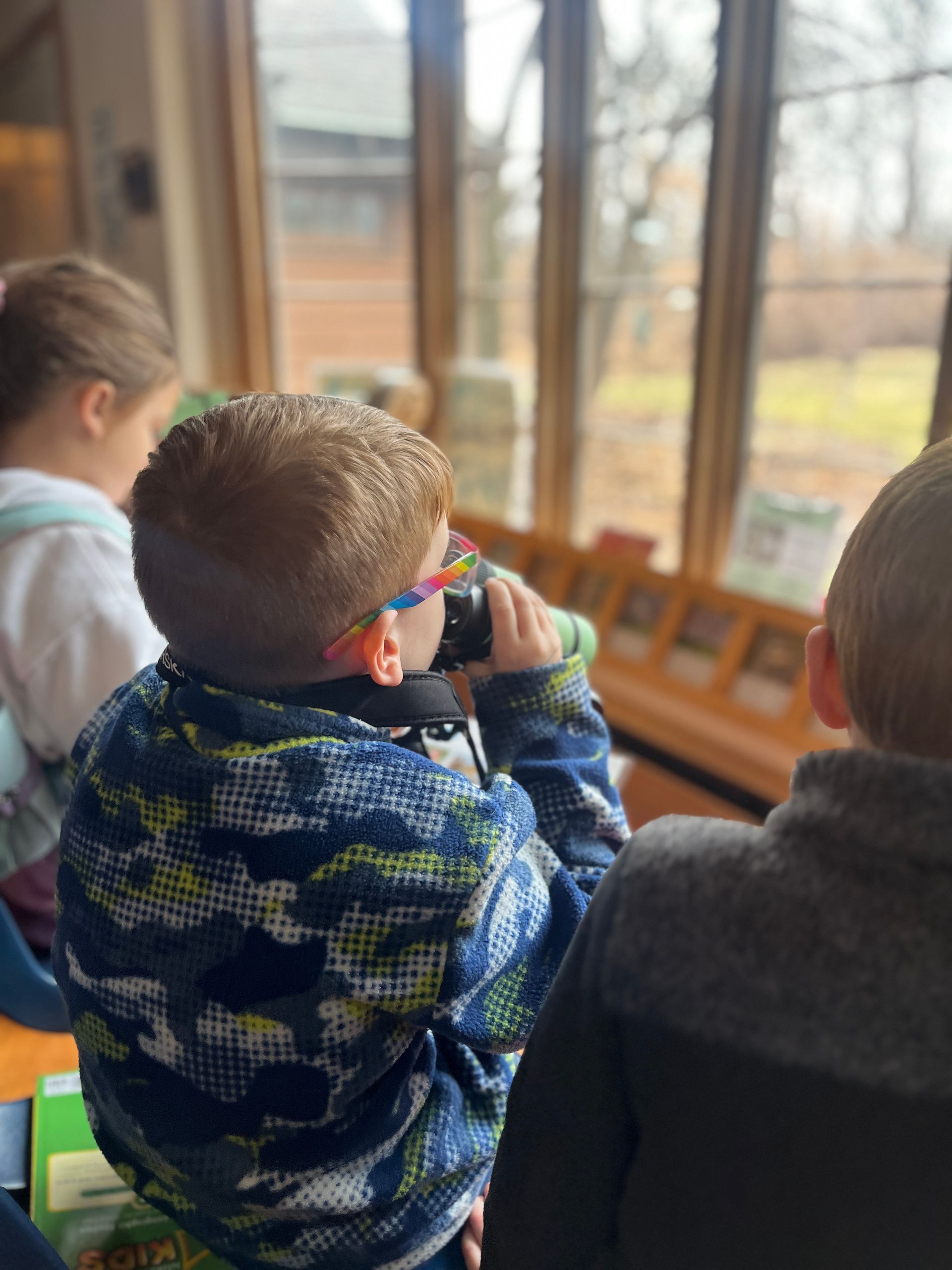 Child with rainbow glasses using binoculars in front of a window.