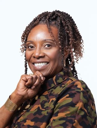 Woman with dark skin smiles, wearing a camouflage shirt, resting a hand on her chin. Braided hair.