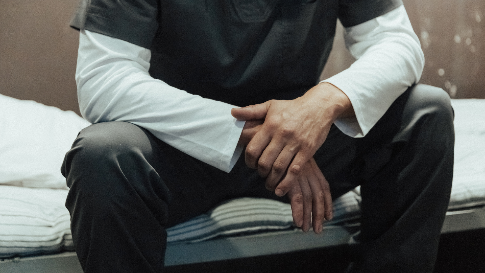 Man in prison uniform sits on a bed, hands clasped. Gray and white clothing, indoor setting.