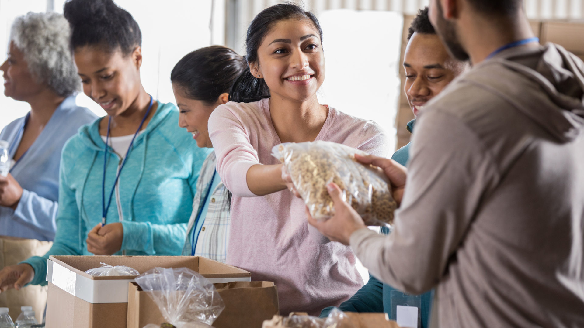 Volunteers packing food, diverse group, smiles, handing out provisions in a warehouse setting.