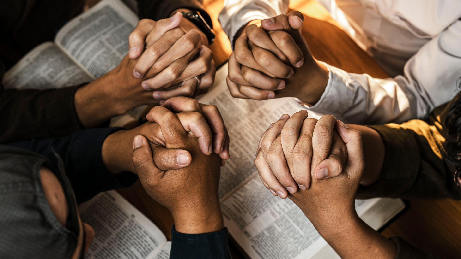Hands clasped in prayer over open books, possibly Bibles. Various skin tones; group setting.