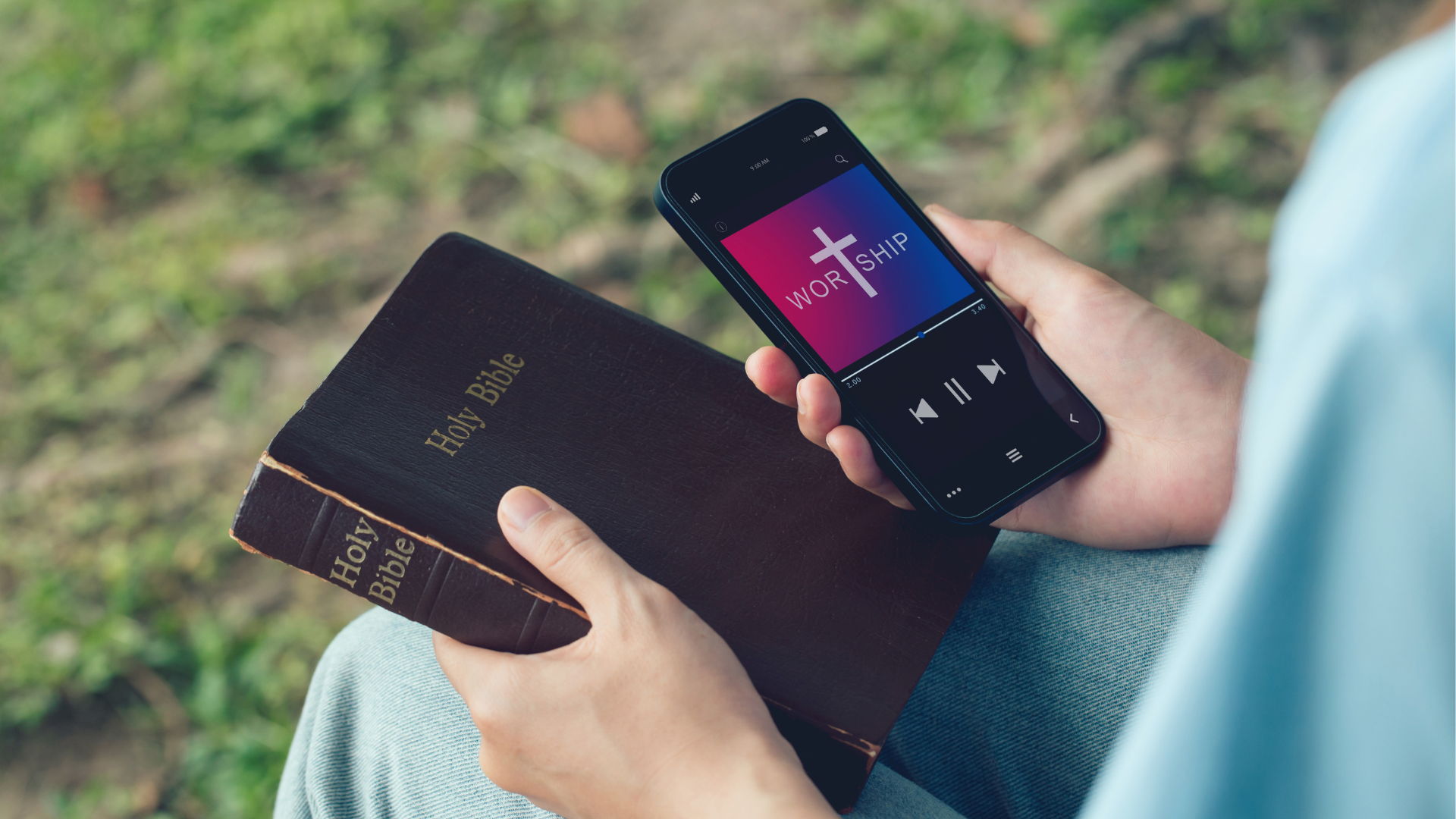 Person holding a Bible and a smartphone playing Christian music outdoors.