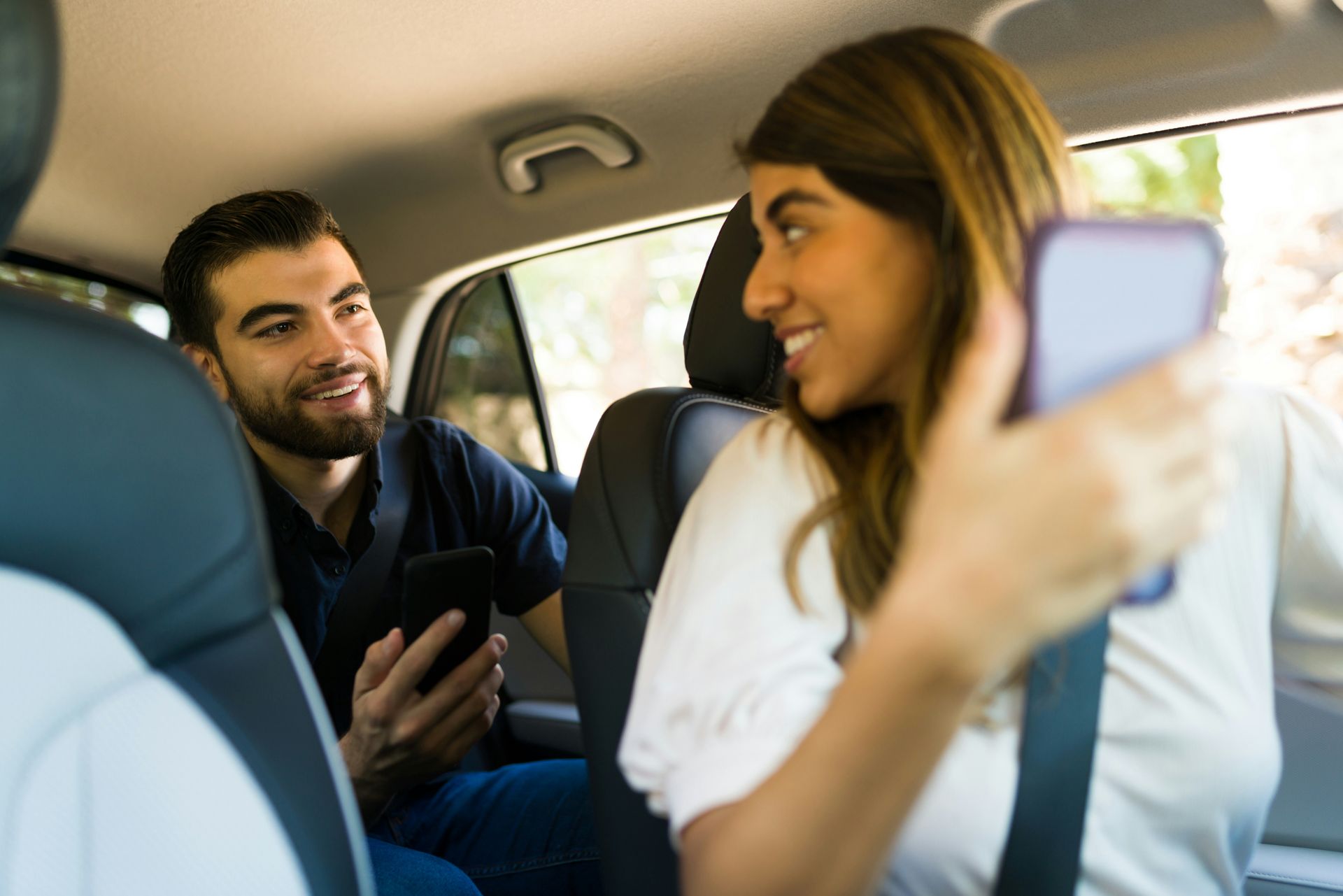 A man and a woman are sitting in the back seat of a car looking at their phones.