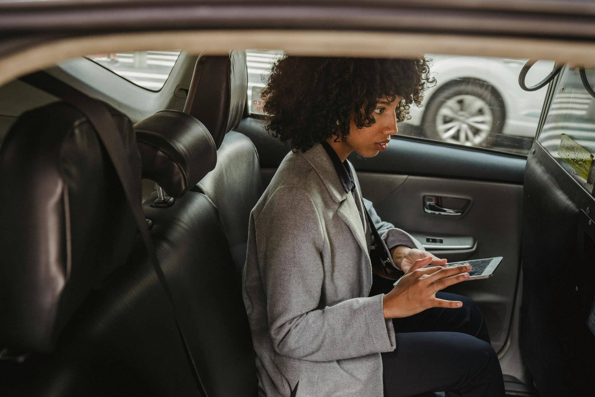 A person with curly hair wearing a gray coat sits in the backseat of a car while using a tablet.