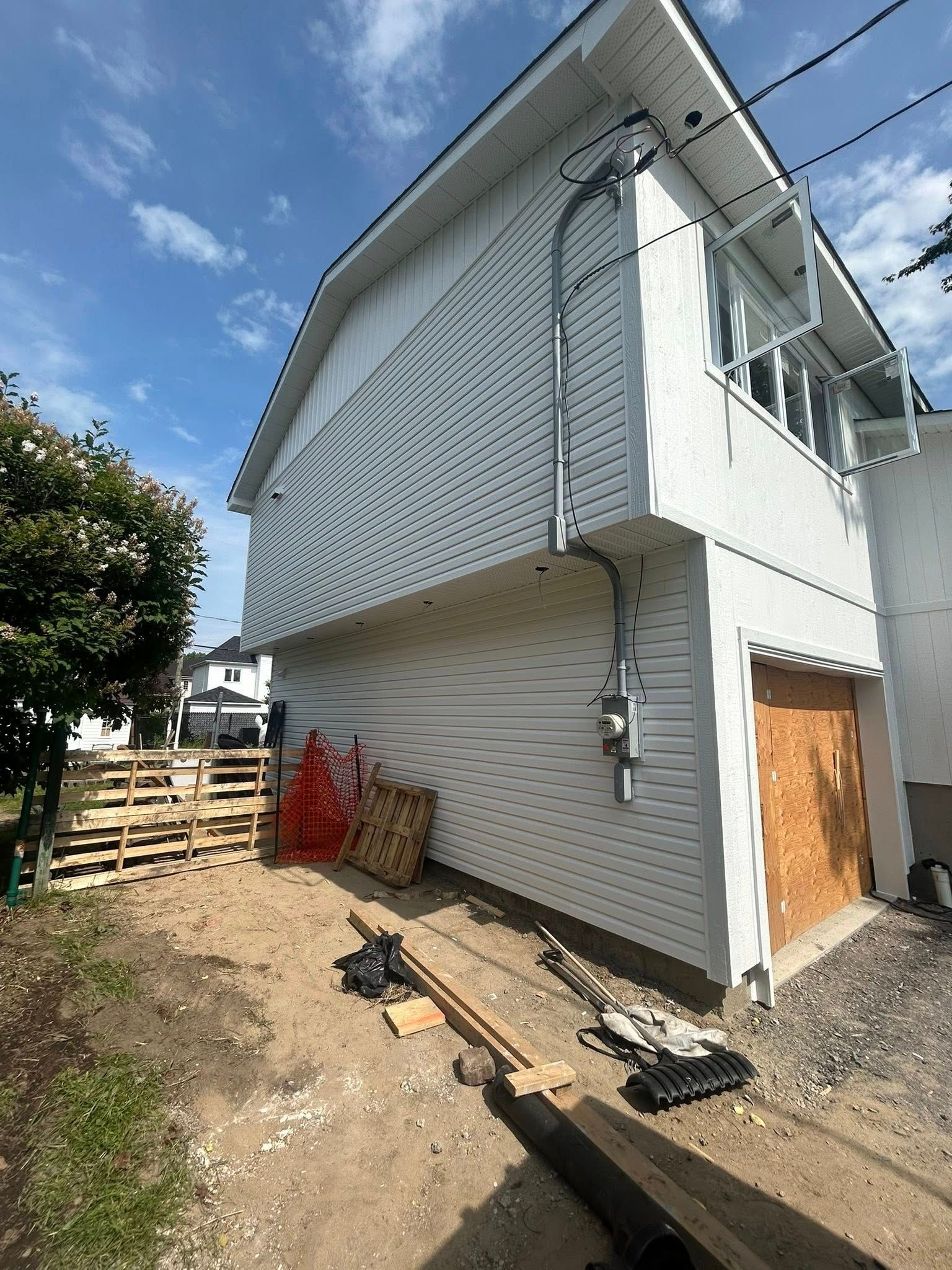 Bâtiment blanc à l'extérieur, avec bardage neuf, conduits électriques et porte en contreplaqué. Matériaux de construction présents.