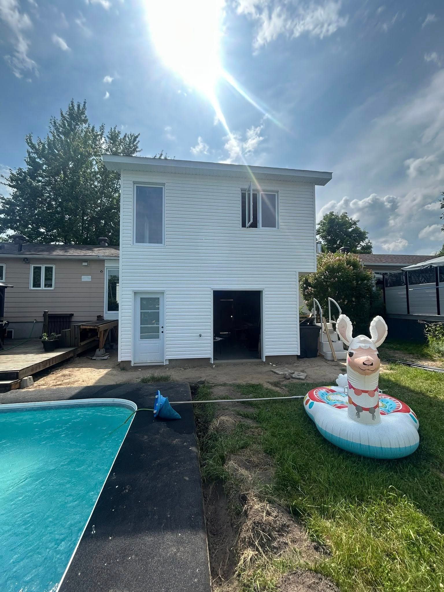 Bâtiment blanc de deux étages avec porte de garage ouverte, à côté d'une piscine. Un lama gonflable flotte sur l'herbe.