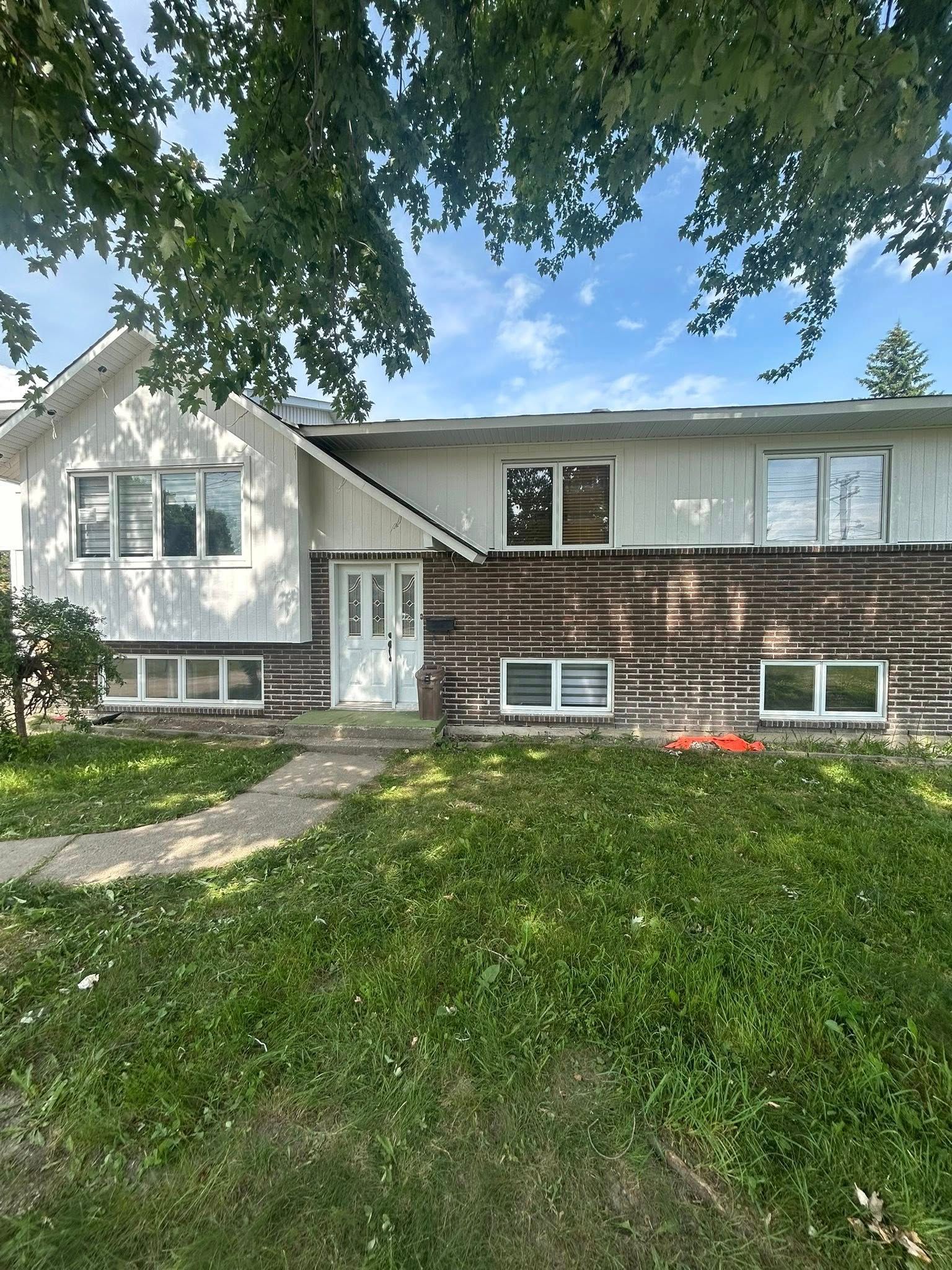 Maison à deux étages avec bardage blanc, façade en briques et pelouse verte sous un ciel nuageux.