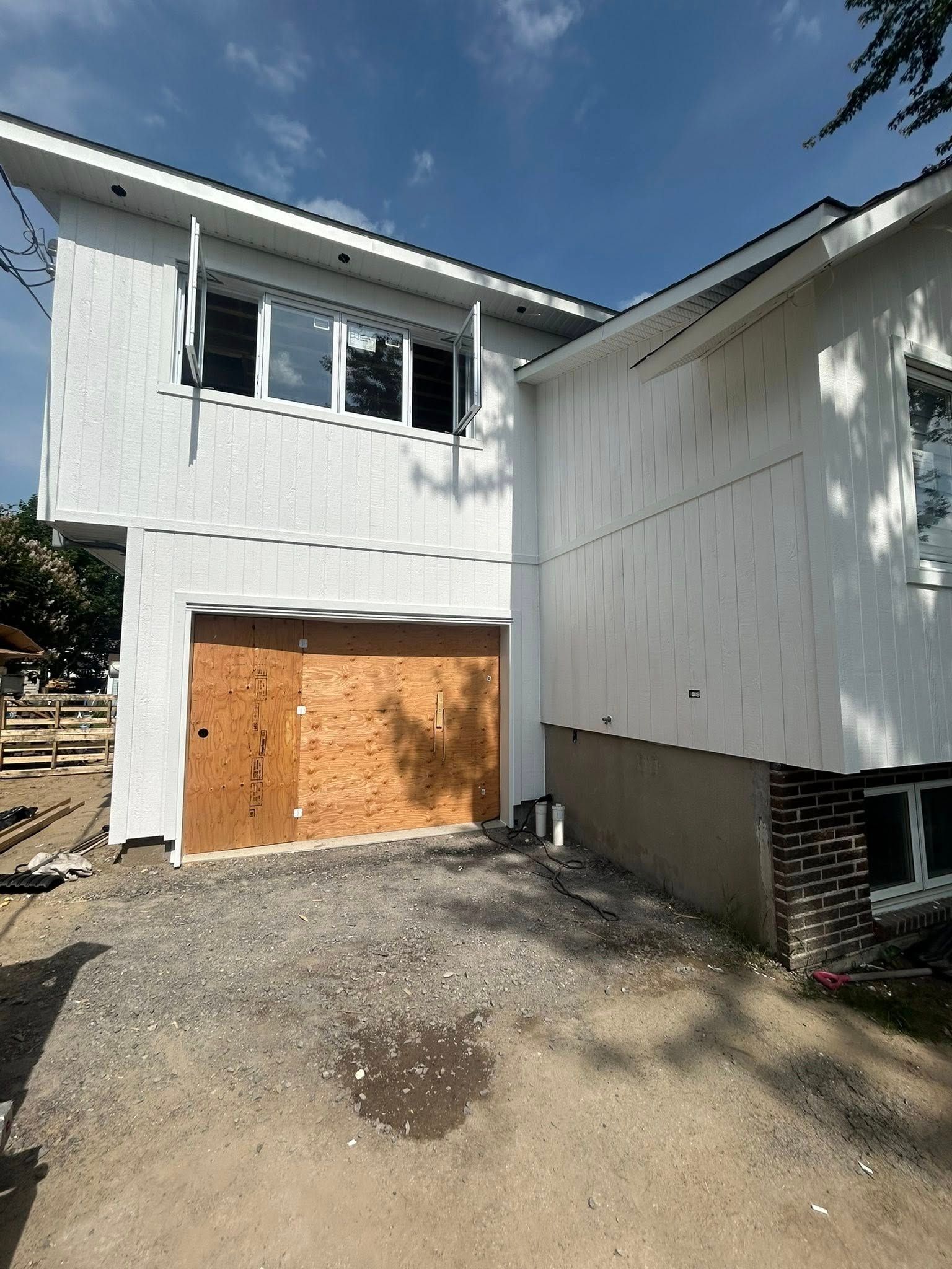 Extérieur d'une maison en construction ; bardage blanc, porte de garage en contreplaqué, allée en gravier.
