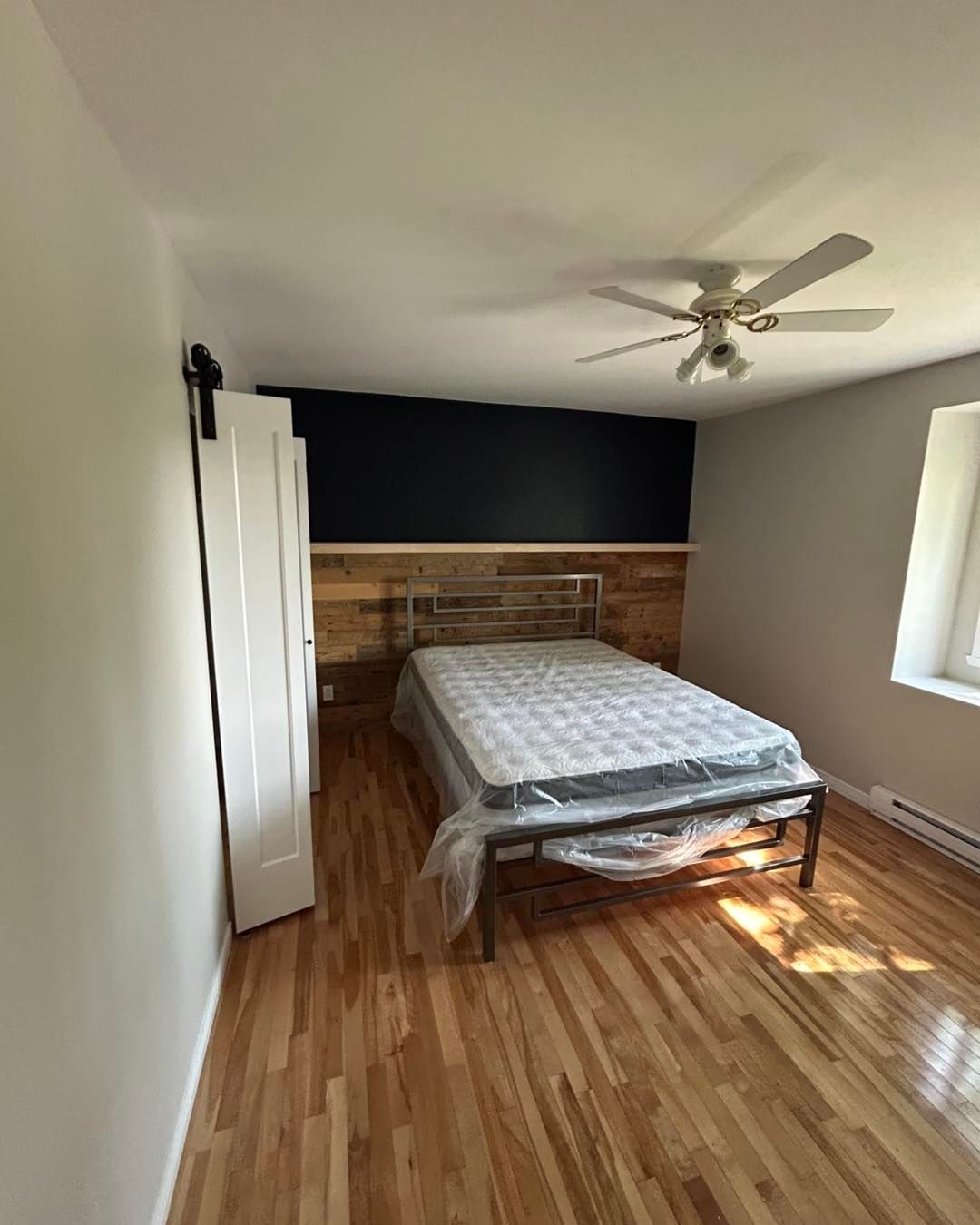 Chambre avec parquet, lit, mur d'accent noir, porte de grange et ventilateur de plafond.